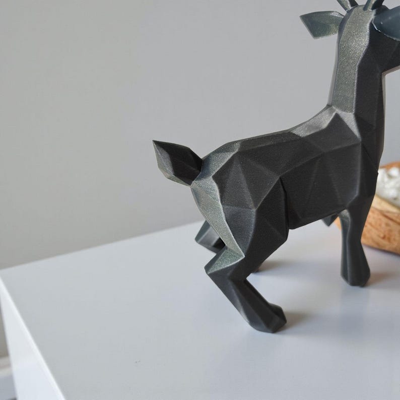 May include: A geometric, black deer figurine with a faceted design. The deer has antlers and is posed in a running stance. The sculpture is made of a dark, matte material and sits on a white surface.