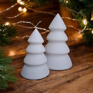 May include: Two white ceramic Christmas tree candles, one larger than the other, on a wooden surface with a string of white lights.