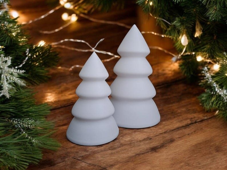 May include: Two white ceramic Christmas tree candles, one larger than the other, on a wooden surface with a string of white lights.