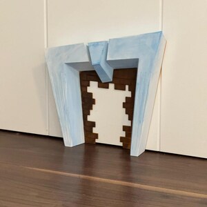 May include: A geometric, abstract sculpture with a light blue painted exterior and a brown and white interior. The sculpture is placed on a dark brown wooden surface, with a white wall in the background. The artwork has a modern, architectural aesthetic.
