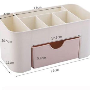 May include: A white and pink organizer with multiple compartments and a drawer. The organizer is 22 cm long, 11 cm wide, and 10.5 cm tall. The drawer is 12.5 cm wide and 5.8 cm tall. The compartments are 13 cm wide and 4 cm deep.