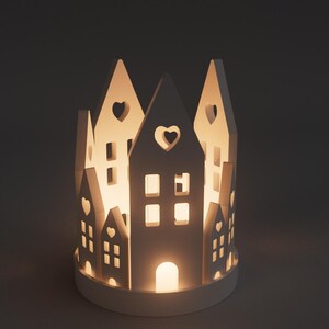 May include: A white ceramic candle holder shaped like a village. The design features cut-out windows and heart-shaped details, with a warm light emanating from within. The base is round and the overall design is minimalist.