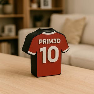 K&ouml;nnte beinhalten: Ein rot-schwarzes Deko-Objekt in Form eines Fu&szlig;balltrikots. Das Trikot hat die Nummer 10 in Wei&szlig; und den Text "PRIM3D" in Wei&szlig;. Das Objekt steht auf einem Holztisch.