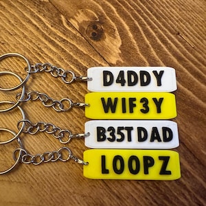 May include: Four keychains with rectangular tags in white and yellow, each with black lettering. The tags read "DADDY", "WIF3Y", "B35T DAD", and "LOOPZ". The keychains have silver-colored rings and chains.