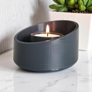 May include: A gray, oval-shaped candle holder with a lit tea light candle inside. The candle holder has a modern design and is made of a solid material. A small succulent plant in a white pot is in the background.