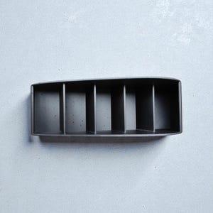 May include: A rectangular, dark gray organizer with six vertical dividers. The container is made of a solid material and is designed to hold small items. The object is set against a light gray background.