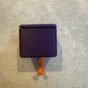 May include: A purple, rectangular hook with a rounded top and an orange, curved hook. The hook is designed to be mounted on a wall or other surface. The hook is made of plastic and is a solid color.