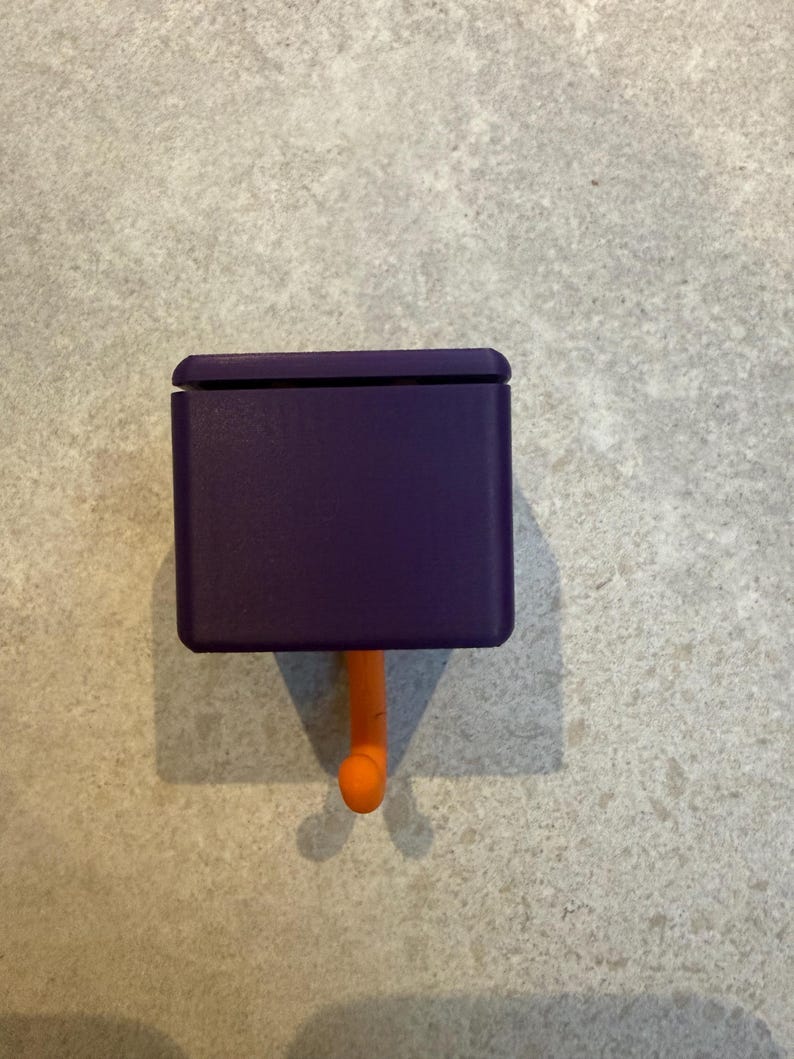 May include: A purple, rectangular hook with a rounded top and an orange, curved hook. The hook is designed to be mounted on a wall or other surface. The hook is made of plastic and is a solid color.