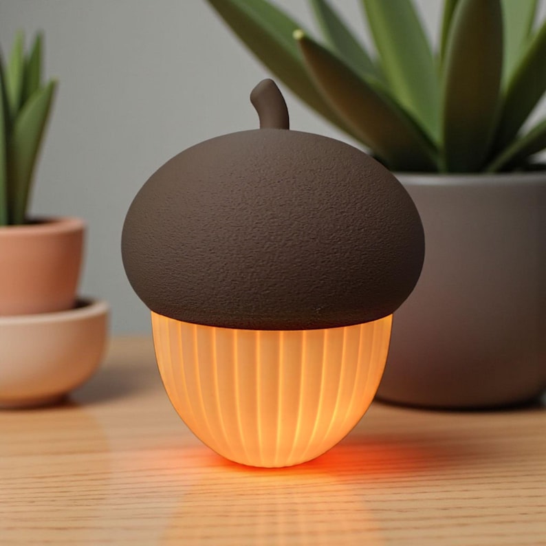 May include: An acorn-shaped lamp with a brown textured top and a ribbed, glowing amber-colored base. The lamp is on a wooden surface, with potted plants in the background. The lamp emits a warm, inviting light.