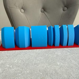 Op de afbeelding: Een set blauwe geometrische vormen, waaronder kubussen en gebogen vormen, gerangschikt op een rode rechthoekige basis. De vormen vari&euml;ren in grootte en ontwerp, waardoor een visueel interessante weergave ontstaat. De objecten zijn gemaakt van een solide materiaal.