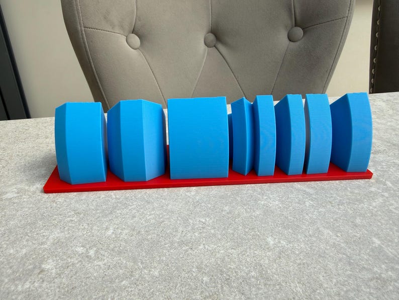 Op de afbeelding: Een set blauwe geometrische vormen, waaronder kubussen en gebogen vormen, gerangschikt op een rode rechthoekige basis. De vormen vari&euml;ren in grootte en ontwerp, waardoor een visueel interessante weergave ontstaat. De objecten zijn gemaakt van een solide materiaal.