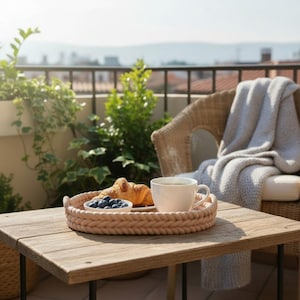 Puede incluir: Una mesa de madera clara en un balc&oacute;n presenta una bandeja tejida con una taza de t&eacute;, un cruas&aacute;n y ar&aacute;ndanos. Una silla de mimbre con una manta gris est&aacute; cerca, con vegetaci&oacute;n en el fondo.