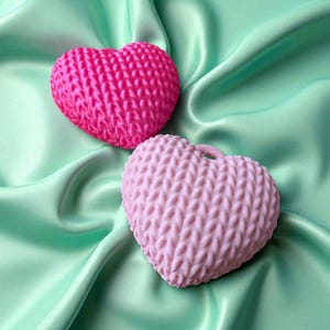 May include: Two heart-shaped items with a knitted texture, one bright pink, the other a pale pink. They are displayed on a mint-green satin fabric, offering a contrast of textures and colours. These appear to be decorative or craft objects.