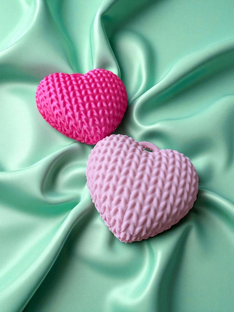 May include: Two heart-shaped items with a knitted texture, one bright pink, the other a pale pink. They are displayed on a mint-green satin fabric, offering a contrast of textures and colours. These appear to be decorative or craft objects.