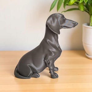 May include: A gray dachshund figurine, sitting upright with its head turned to the side. The sculpture is made of a smooth material and has detailed features. The dog statue is placed on a wooden surface.
