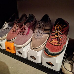 May include: A collection of shoes of various styles and colors, including blue, red, brown, and grey sneakers, resting on white and orange shoe organizers. The shoes are arranged on a black shelf.