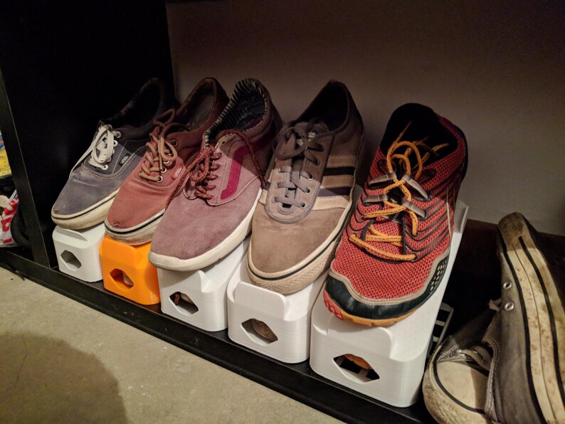 May include: A collection of shoes of various styles and colors, including blue, red, brown, and grey sneakers, resting on white and orange shoe organizers. The shoes are arranged on a black shelf.