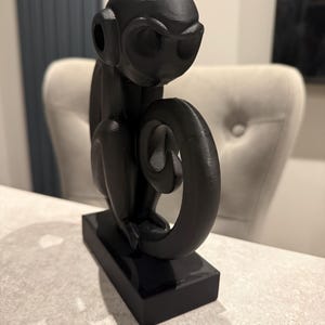 Puede incluir: Una figura de mono negro con una cola enroscada, sentada sobre una base rectangular. El mono tiene orejas grandes y est&aacute; sentado. La figura est&aacute; hecha de un material liso y brillante.