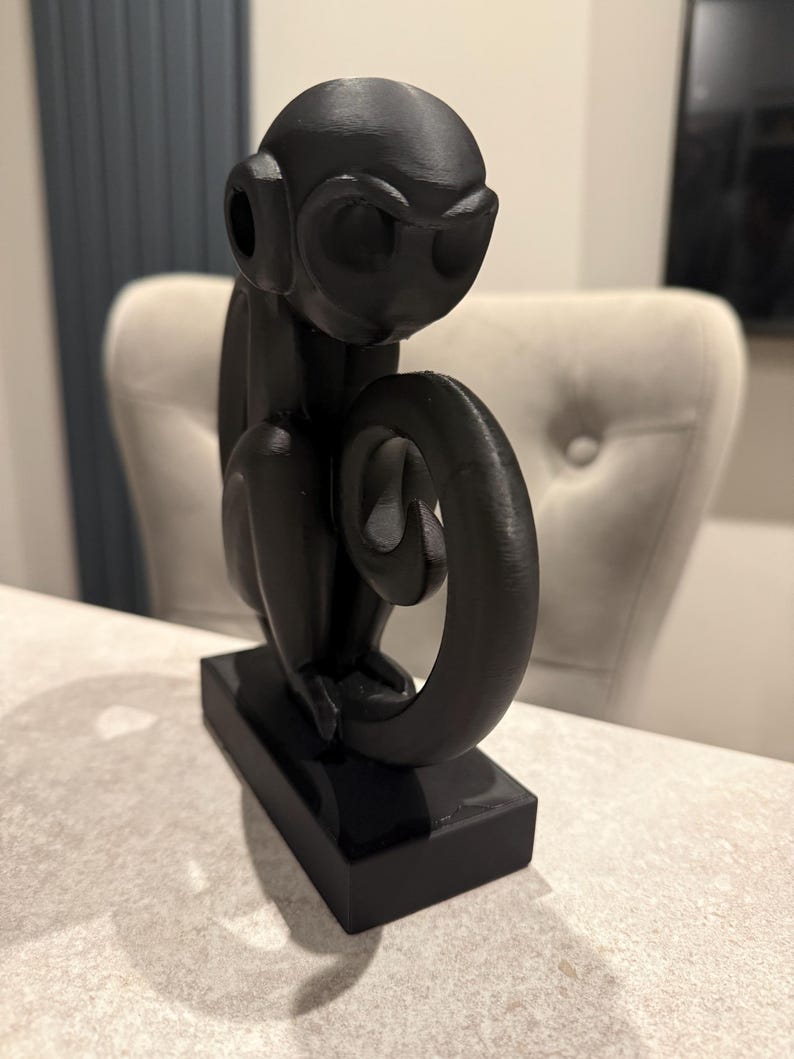 Puede incluir: Una figura de mono negro con una cola enroscada, sentada sobre una base rectangular. El mono tiene orejas grandes y est&aacute; sentado. La figura est&aacute; hecha de un material liso y brillante.