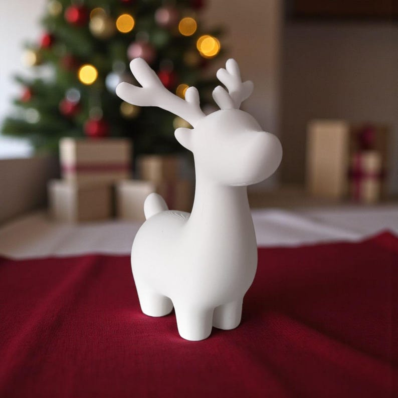 May include: A white ceramic reindeer figurine with antlers, standing on a red surface. The reindeer has a smooth, matte finish and a stylized design. Christmas tree and wrapped gifts are in the background.
