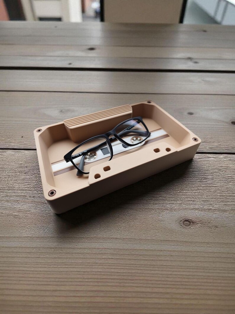 Pu&ograve; includere: Un vassoio beige a forma di cassetta con una striscia bianca e occhiali con montatura nera. Il vassoio ha una piccola sezione rialzata su un lato. Gli occhiali sono neri. Il vassoio &egrave; su una superficie di legno.