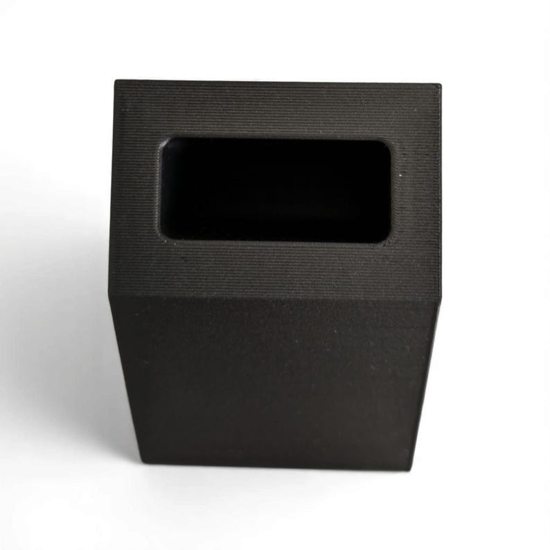 May include: A black, rectangular, open-top container with a textured surface. The container has a rectangular opening at the top and a trapezoidal base. The object is set against a white background.