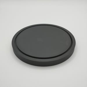 May include: A round, black coaster with a slightly raised edge. The coaster is made of a solid, matte material and is designed to protect surfaces from drink condensation. The simple design is suitable for various home decor styles.