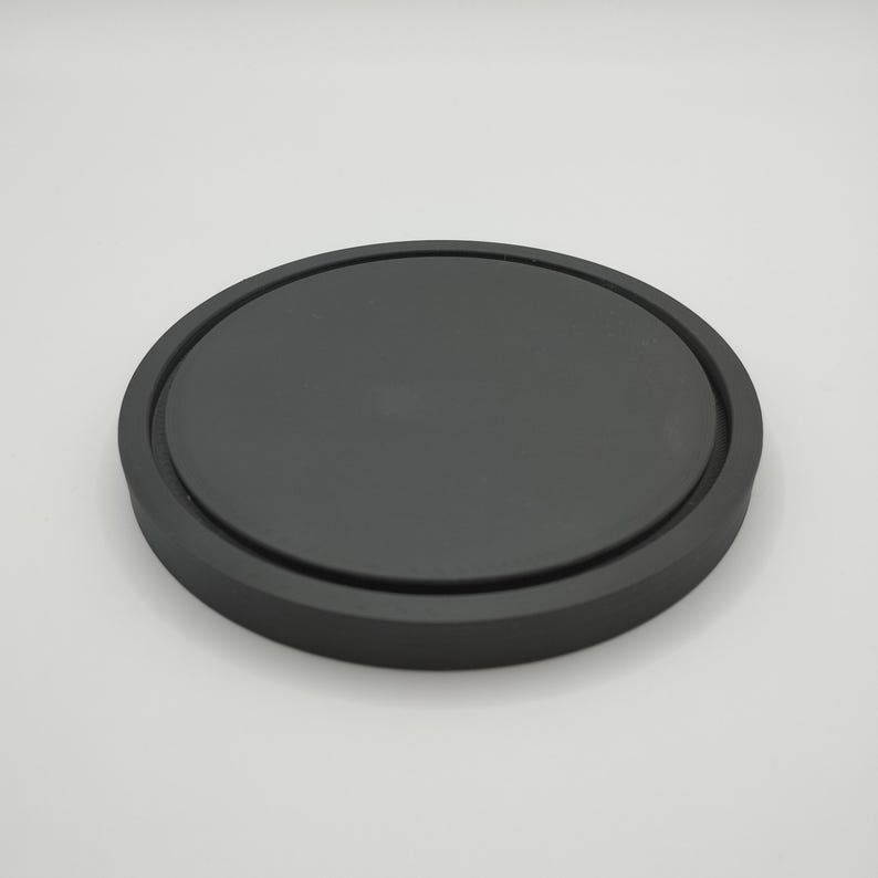 May include: A round, black coaster with a slightly raised edge. The coaster is made of a solid, matte material and is designed to protect surfaces from drink condensation. The simple design is suitable for various home decor styles.