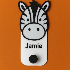 May include: A white and black zebra-themed nameplate with the name "Jamie" printed in black. The nameplate has a black circular knob at the bottom and is set against an orange background. The zebra's head is at the top.
