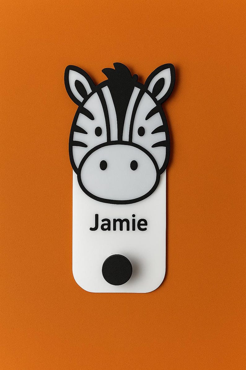 May include: A white and black zebra-themed nameplate with the name "Jamie" printed in black. The nameplate has a black circular knob at the bottom and is set against an orange background. The zebra's head is at the top.