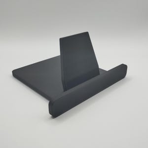 May include: A dark grey, minimalist phone or tablet stand. The stand has a rectangular base with a raised back and a front lip to hold a device. The back piece is angled for optimal viewing. The stand is made of a smooth, solid material.