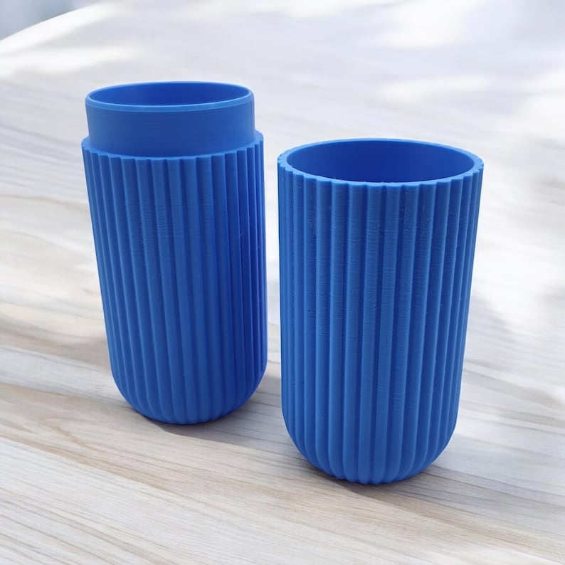 May include: Two blue cylindrical containers with vertical ribbed detailing. One container has a lid, and the other is open. The containers are made of a solid blue material and sit on a light-colored wooden surface.