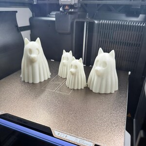 Pu&ograve; includere: Quattro figure di cani bianchi stampate in 3D, di dimensioni variabili, sono disposte su una superficie metallica all'interno di una stampante 3D. Le figure hanno un aspetto spettrale con forme drappeggiate e facce di cane. La stampante ha un'etichetta Bambu Lab.