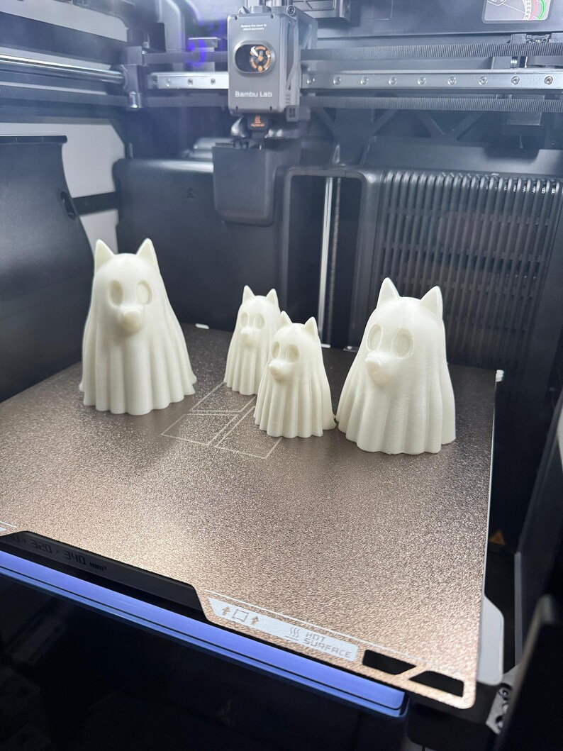 Pu&ograve; includere: Quattro figure di cani bianchi stampate in 3D, di dimensioni variabili, sono disposte su una superficie metallica all'interno di una stampante 3D. Le figure hanno un aspetto spettrale con forme drappeggiate e facce di cane. La stampante ha un'etichetta Bambu Lab.