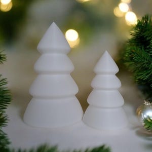 May include: Two white ceramic Christmas tree ornaments, one larger than the other, with a rounded, layered design. The ornaments are on a white surface with green pine branches in the background.
