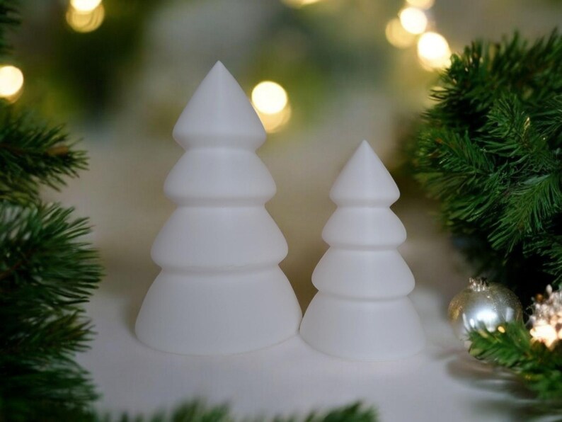 May include: Two white ceramic Christmas tree ornaments, one larger than the other, with a rounded, layered design. The ornaments are on a white surface with green pine branches in the background.