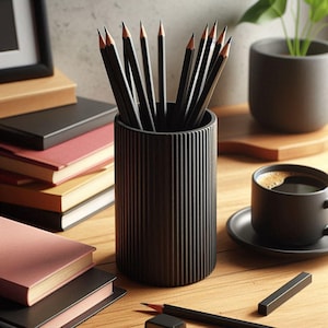 Op de afbeelding: Een zwarte potloodhouder met verticale groeven, gevuld met geslepen potloden. Een kopje koffie, boeken en een kleine plant in een pot staan ook op het houten oppervlak. De potloden hebben een zwarte body en een roze gum.