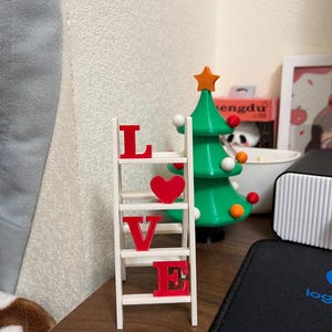 May include: A white wooden ladder shelf with the word "LOVE" spelled out in red letters and a red heart. A green Christmas tree with orange and white ornaments is in the background.