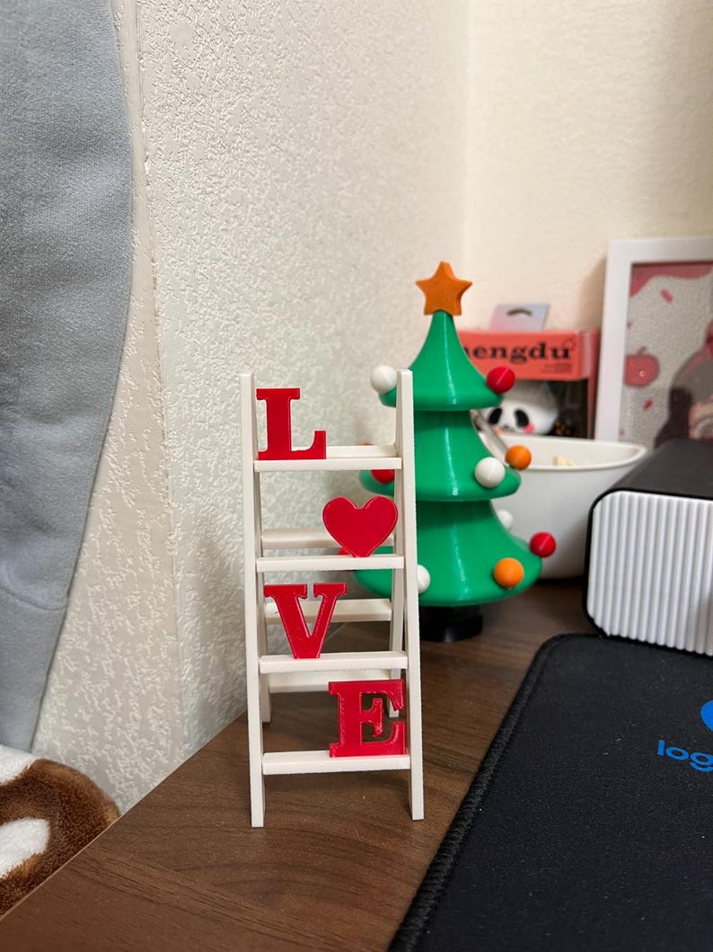 May include: A white wooden ladder shelf with the word "LOVE" spelled out in red letters and a red heart. A green Christmas tree with orange and white ornaments is in the background.