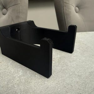 Op de afbeelding: Een zwart, 3D-geprint object met een rechthoekige basis en twee rechtopstaande steunen. Het object heeft een gebogen bovenkant en een gat in de buurt van de bovenkant. De steunen zijn schuin en hebben een gekerfd ontwerp. Het object ligt op een lichtgrijze ondergrond.