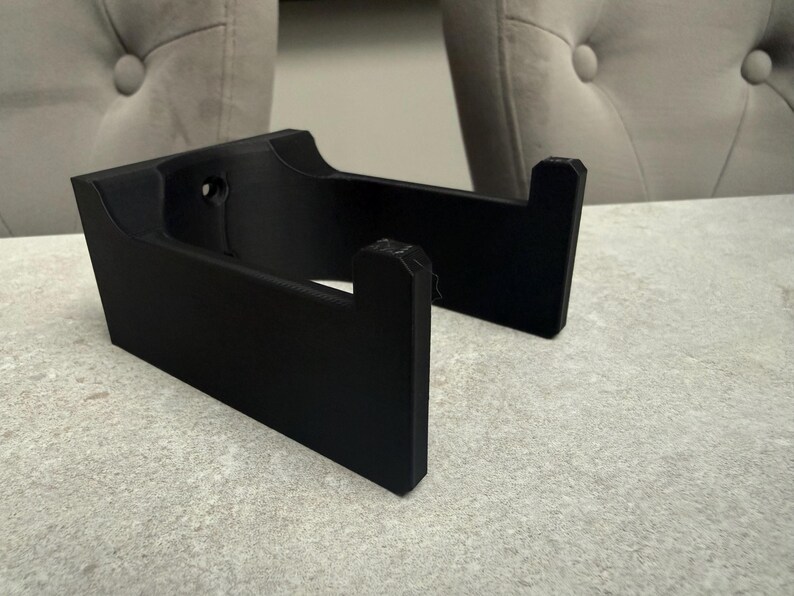 Op de afbeelding: Een zwart, 3D-geprint object met een rechthoekige basis en twee rechtopstaande steunen. Het object heeft een gebogen bovenkant en een gat in de buurt van de bovenkant. De steunen zijn schuin en hebben een gekerfd ontwerp. Het object ligt op een lichtgrijze ondergrond.