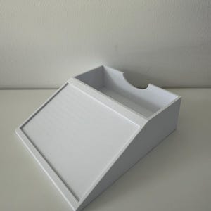 May include: A white, angled desk organiser with a textured, sloped front and a rectangular top compartment. The organiser has a curved cutout on one side, designed to hold papers or office supplies.