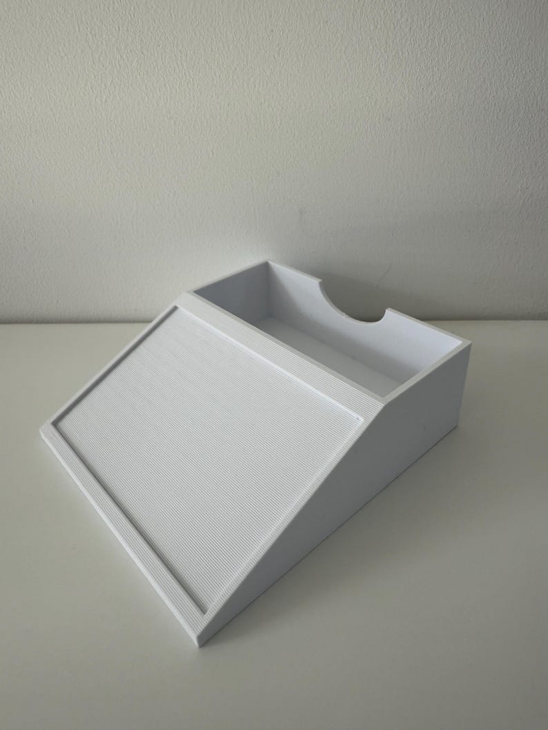 May include: A white, angled desk organiser with a textured, sloped front and a rectangular top compartment. The organiser has a curved cutout on one side, designed to hold papers or office supplies.