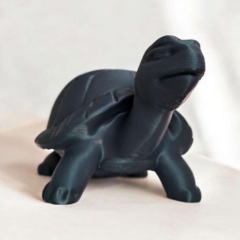 Puede incluir: Figura de tortuga gris oscuro con caparaz&oacute;n y cabeza detallados. La escultura est&aacute; hecha de un material liso y tiene una superficie ligeramente reflectante. La tortuga est&aacute; posada con la cabeza levantada.