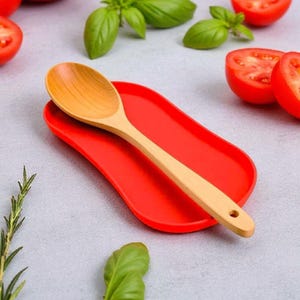 May include: A wooden spoon sits on a vibrant red spoon rest. The spoon is light brown with a rounded bowl and a long handle. The spoon rest is a solid, curved shape. Fresh basil and halved tomatoes are in the background.