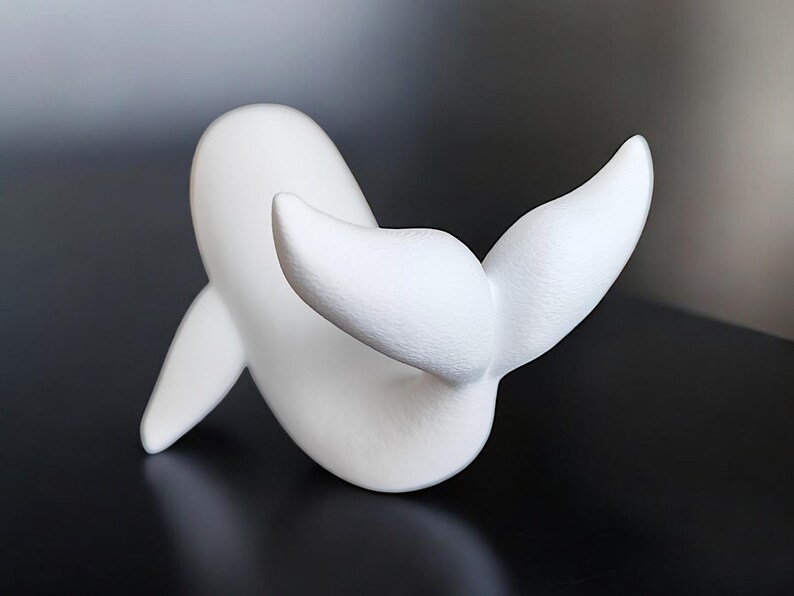May include: A white whale sculpture with a smooth, matte finish. The whale is depicted in a playful pose, with its tail flukes raised. The sculpture is a minimalist design, perfect for home decor.