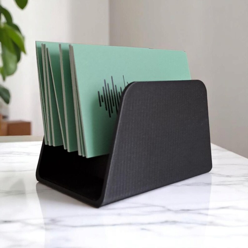 May include: A black desktop organizer holding several mint green cards. Each card features a black sound wave design. The organizer is made of a matte black material and sits on a white marble surface.