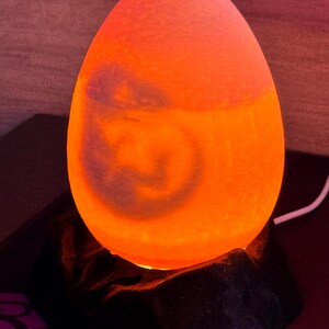May include: An egg-shaped lamp with a warm orange glow. The lamp sits on a black, textured base. The light emanates from within, creating a soft, ambient illumination. A decorative accent for any room.