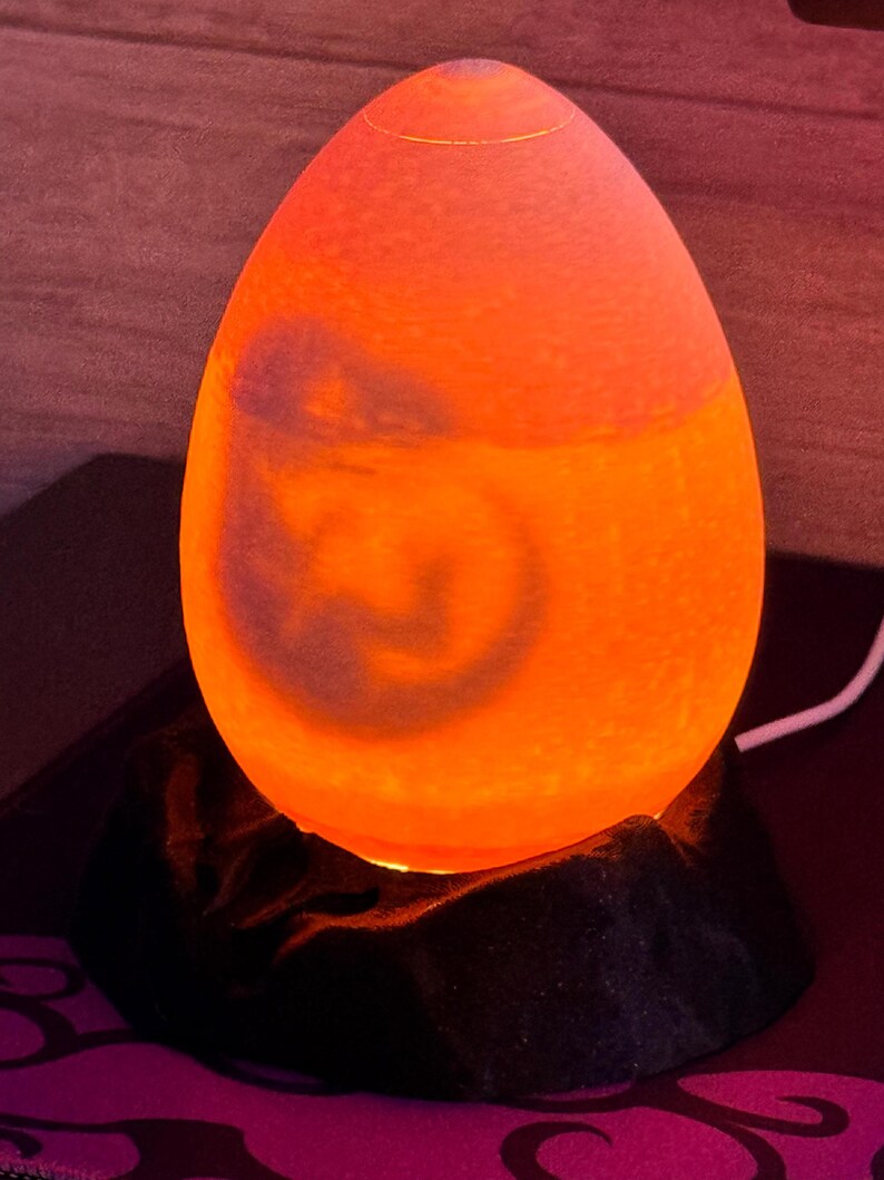 May include: An egg-shaped lamp with a warm orange glow. The lamp sits on a black, textured base. The light emanates from within, creating a soft, ambient illumination. A decorative accent for any room.