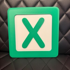 May include: A square, green-framed object with a white center featuring a large, green "X". The object is likely a game piece or a decorative item. The design is simple and bold, with a clear contrast between the green and white elements.